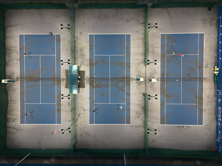 An aerial view of three outdoor tennis courts with blue playing surfaces and white line markings, situated within a fenced sports complex. The courts are separated by low green barriers and have a grey surrounding pavement area. The surfaces show some dirt and scattered debris, indicating recent use or exposure to the elements. On the left court, there is a small black maintenance or cleaning device, while the middle court features a table tennis table with black surface and white borders, positioned near the net area. The right court has a small cluster of tennis balls and a few scattered items, possibly sports equipment, near the service line. The environment appears well-lit with natural daylight, and the overall scene captures a sports facility that may require cleaning and rubbish removal, something that services like Waste Removal Roehampton could support in maintaining a tidy environment for recreational use.