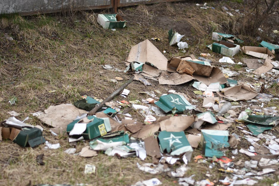 An outdoor scene showing a混乱 of discarded cardboard boxes, some flattened and others torn, with visible green and white branding, scattered across a patch of uneven grassy ground near a fence. The boxes are partially crushed, with layers of paper, torn edges, and packaging debris mixed in. Among the waste are various pieces of paper and plastic wrappers, some crumpled or torn, contributing to an overall messy appearance. The ground features patches of brown soil and sparse grass, indicating an unmanaged area prone to littering. In the background, a weathered metal fence marks the boundary of the site, with dry, leafless shrubs and small plants growing nearby. Natural lighting illuminates the scene, highlighting the textures of the cardboard and litter, typical of a site where private rubbish clearance, such as through Waste Removal Roehampton, might be required to tidy up and manage waste outside of official disposal channels.