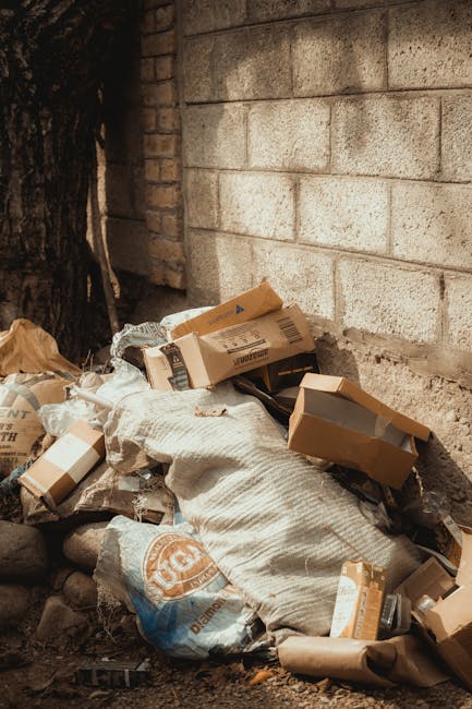 A pile of discarded cardboard boxes, some flattened and some partially assembled, along with plastic packaging and a woven fabric sack, all resting against a brick wall outdoors. The boxes vary in size and are light brown with printed labels or barcodes visible on some. The fabric sack appears to be beige or off-white, with a coarse texture. The scene is set on uneven ground with scattered small rocks or debris, adjacent to a textured brick wall and a large tree trunk on the left side of the image. Natural daylight illuminates the scene, casting soft shadows. The arrangement of waste objects suggests an informal or on-site rubbish collection, typical of private waste disposal or clearance services, such as those offered by Waste Removal Roehampton. The environment indicates a non-residential area, possibly a back alley, garden boundary, or service access point, where waste is temporarily accumulated before removal or disposal.