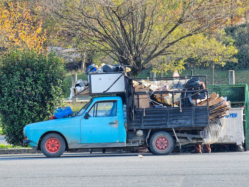 A small, blue, vintage pickup truck parked on the side of a street with its cargo area on the flatbed filled with various discarded items including cardboard boxes, black plastic bags, and miscellaneous debris. The truck shows signs of wear with faded paint and red steel wheels, and is positioned next to a curb beneath a large tree with predominantly green and some yellow leaves. In the background, there is a metal fence and additional greenery, indicating a suburban or park-like environment. This scene appears to depict a private waste collection or rubbish removal process, possibly managed by Waste Removal Roehampton, using an alternative or independent method of rubbish disposal outside of local authority services.