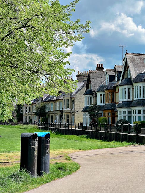 A row of old terraced houses with bay windows and brick facades on a residential street, featuring varied rooflines with chimneys and dormer windows under a partly cloudy sky. A large leafy tree with bright green foliage extends its branches across the upper left of the image, providing partial shade for the street below. In the foreground, a paved footpath runs past two black cylindrical rubbish bins positioned on a grassy verge, which appears to be part of a public park or green space. Behind the bins, a low wooden fence separates the grassy area from the pavement and houses. This scene captures a quiet suburban setting during daytime, with natural light illuminating the buildings and greenery, reflecting typical UK residential environments that may require routine rubbish collection or private waste handling services, such as those offered by Waste Removal Roehampton.